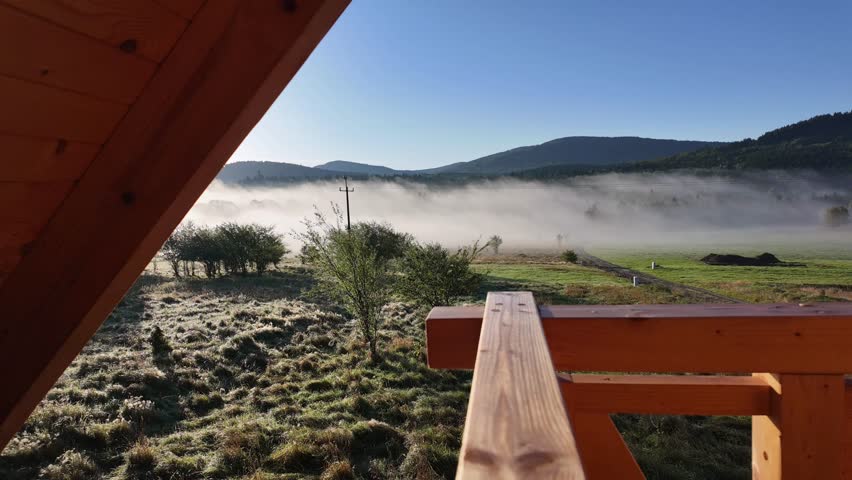 Balcony view of meadow, forest, and mountains on a frosty sunny morning. Narrow mist strips drift left, highlighting serene natural winter landscape.