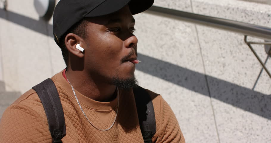 African American man chewing on outdoor steps with earbuds, turning toward sunlight and smiling. Urban, casual, relaxed, sunlight, contemplative, outdoor, modern
