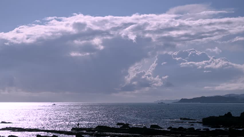 Wide shot of white clouds moving over the sparkling ocean at dusk in Wanli District New Taipei City Taiwan A lone person stands on the dark rocky shore fishing.