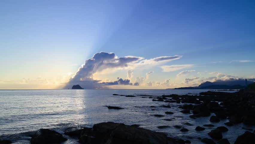 Dramatic wide shot of beautiful sun rays shooting into the colorful blue and purple sky over Keelung Islet and the ocean during pre sunrise on the rocky coast of Wanli Taiwan.