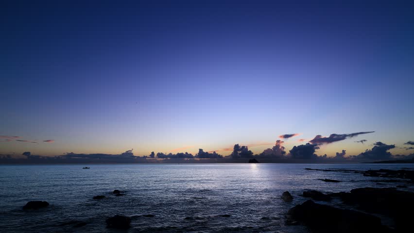 Dawn light fans above Keelung Islet, seen from Wanli District in New Taipei. Crepuscular rays rise over calm sea and dark coastal rocks.
