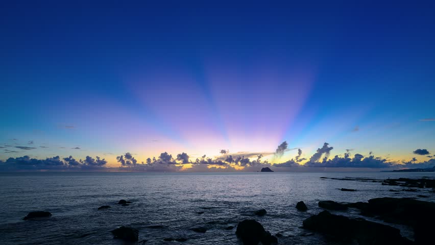 Dramatic wide shot of beautiful sun rays shooting into the colorful blue and purple sky over Keelung Islet and the ocean during pre sunrise on the rocky coast of Wanli Taiwan.