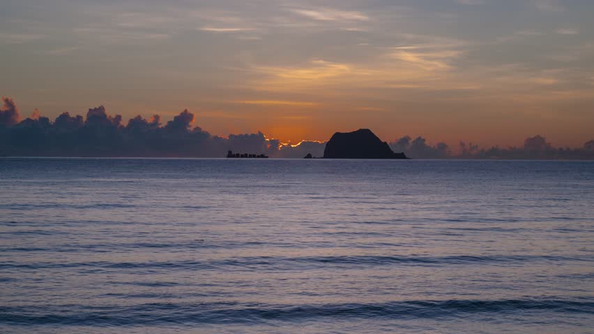 Scenic wide shot of the bright orange and golden sun slowly rising behind the silhouette of Keelung Islet and clouds on the horizon in Wanli District New Taipei City Taiwan.