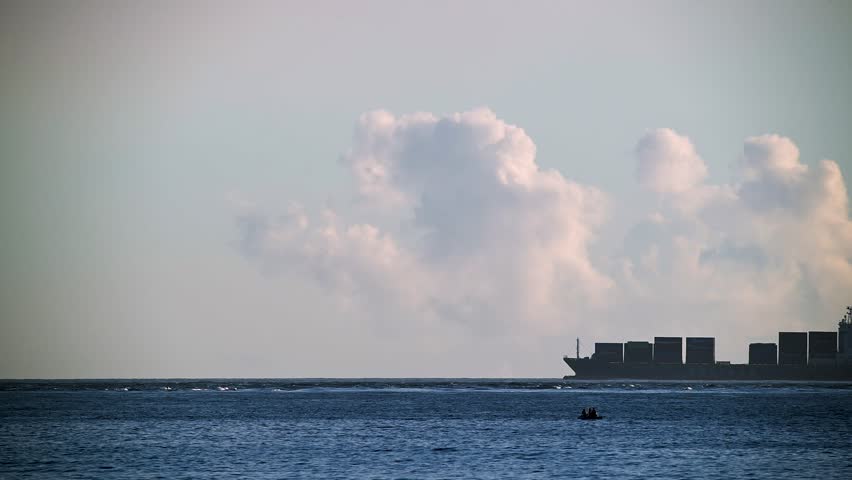 Medium shot of a large container ship moving slowly on the vast ocean horizon under bright white cumulus clouds in Wanli District New Taipei City Taiwan during morning time
