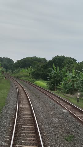 Double railway tracks stretching into the distance through lush tropical greenery under a cloudy sky. Scenic Indonesian train travel perspective.
