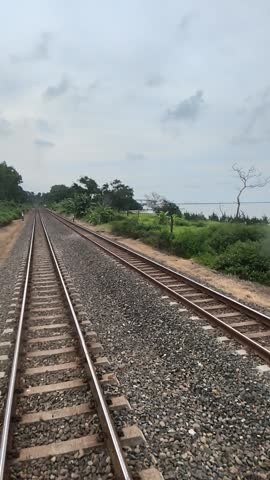 Double railway tracks stretching into the distance through lush tropical greenery under a cloudy sky. Scenic Indonesian train travel perspective.