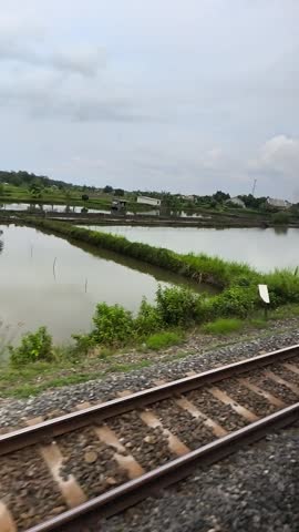 Double railway tracks stretching into the distance through lush tropical greenery under a cloudy sky. Scenic Indonesian train travel perspective.