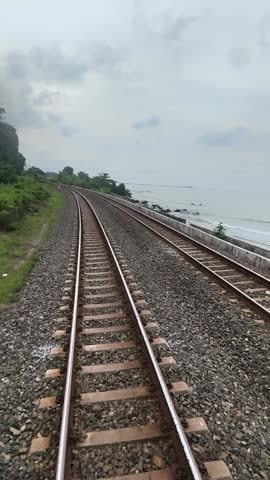 Double railway tracks stretching into the distance through lush tropical greenery under a cloudy sky. Scenic Indonesian train travel perspective.
