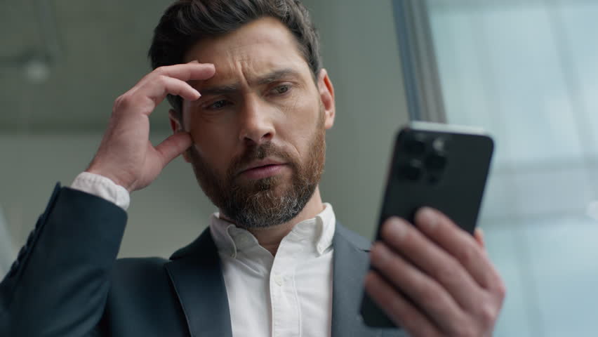 Shocked confused annoyed Caucasian male guy businessman business man in office looking smartphone reading bad news upset with mobile phone frustrated stressed failure app sad negative work problem