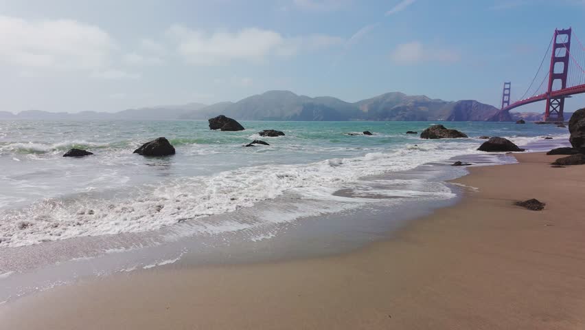 Waves roll onto sandy shore at Baker Beach with Golden Gate Bridge in background, San Francisco, California, USA. Iconic coastal view with ocean, rocks and Marin Headlands