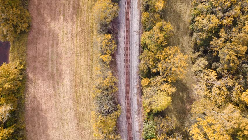 Aerial view of train tracks along a river cutting between open field and wooded trees in early autumn.