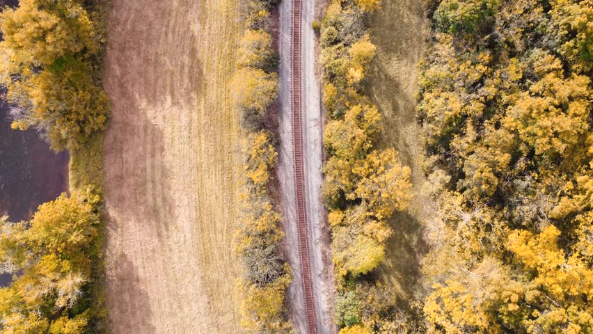 Aerial view of train tracks along a river cutting between open field and wooded trees in early autumn.
