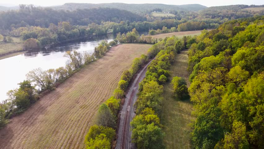 Aerial view of train tracks along a river cutting between open field and wooded trees in early autumn.