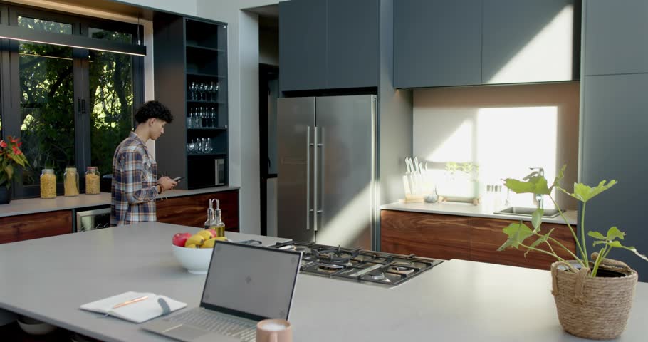 Man using phone in kitchen showing AR causing emojis emerging from right, growing, filling space. Person, remote, workspace, smartphone, laptop, daylight, home