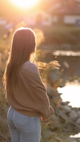 Young woman standing by the river on a cold autumn morning, wrapping herself in a warm sweater. Peaceful cozy atmosphere, golden sunlight, and gentle breeze.