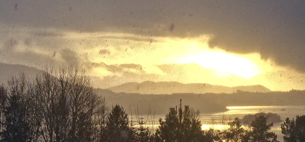Short video clip capturing a heavy snowfall or sleet against the backdrop of a dramatic sunset over a mountain range and a reflecting body of water, creating a golden, grainy effect.