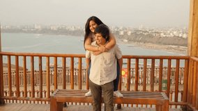 Young man giving his girlfriend a piggyback ride on a wooden promenade. The happy couple is smiling and laughing with a beautiful sea and cityscape background - Powered by Shutterstock - Get 15% off with code: PIKWIZARD15