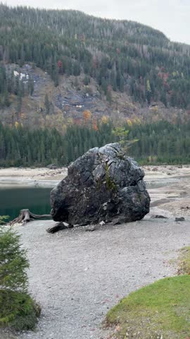 The scene of Austrian Alps with a big stone and tree stumps on the sandy shore of Gosau lake (Upper Gosausee) with forested hills across the lake. Austria. 