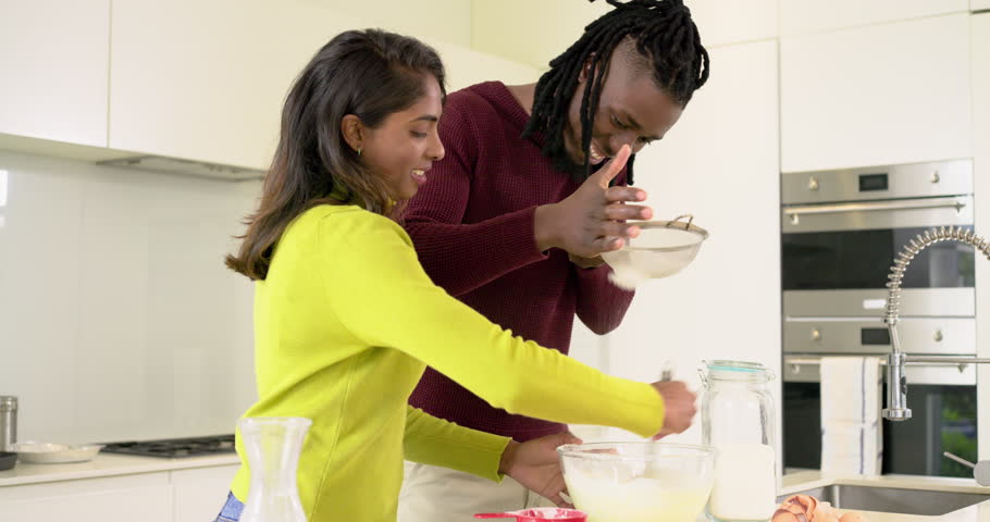 Diverse couple whisking batter, tapping sieve, sifting flour, tasting mixture at kitchen island. Marriage, collaboration, warmth, intimacy, domestic, lifestyle, bright