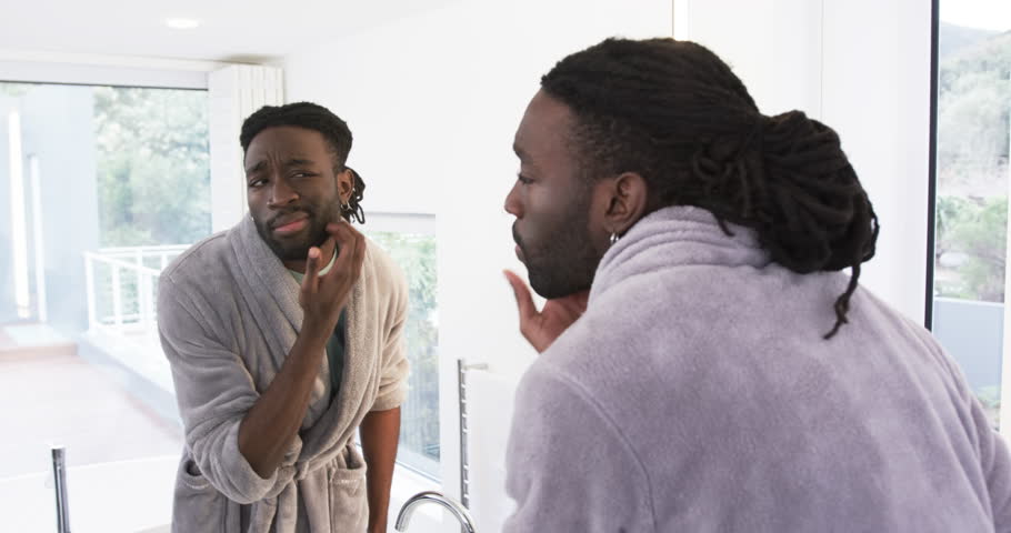 African American man in late 20s entering bathroom in robe checking beard rubbing chin in mirror. Grooming, skincare, self-care, vanity, reflection, masculinity, grooming routine