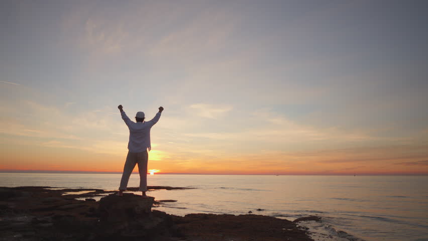 A person stands on a rocky shore at sunset, arms raised in triumph. The serene ocean and vibrant sky evoke mindfulness and reflection, symbolizing aspirations.