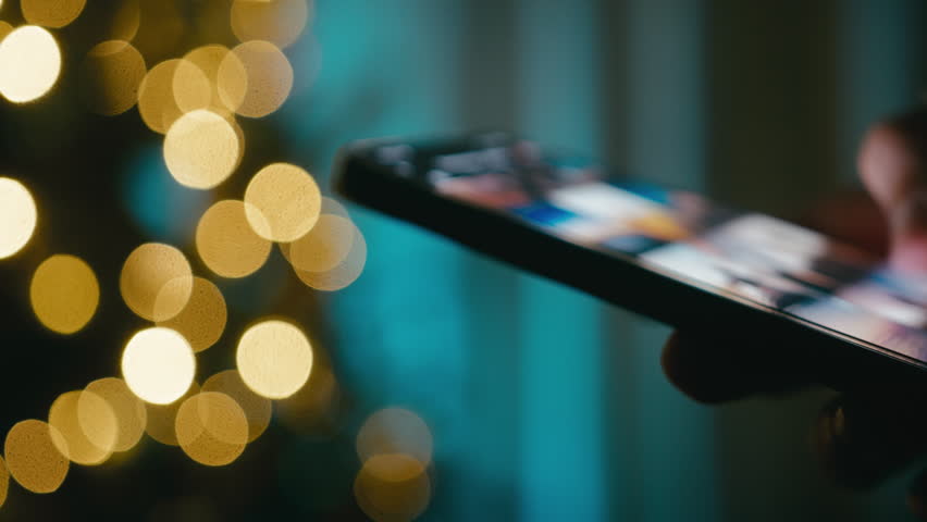Hand flips through photos on a smartphone at night in front of a Christmas tree