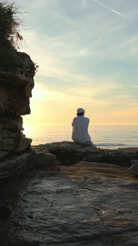 A solitary figure sits on rocky terrain, facing the serene ocean at sunset. The warm glow of the sun creates a peaceful ambiance, evoking mindfulness and reflection.