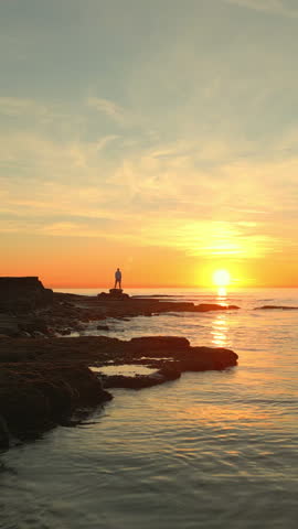 Two figures on a rocky shore at sunset, arms outstretched, embody mindfulness and reflection. The serene ocean and golden sky evoke tranquility and escape, promoting mental wellbeing and aspirations.