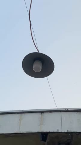 A simple street lamp hanging by a wire beneath a roof line against a clear, soft blue sky background.