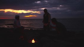 Three silhouetted figures enjoy drinks and conversation by a beach bonfire at sunset. The serene ocean and vibrant sky evoke themes of mindfulness, reflection, and escaping the daily grind. - Powered by Shutterstock - Get 15% off with code: PIKWIZARD15