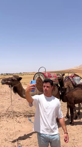 Young athletic muscled tourist male taking a selfie and pictures with his smartphone of a camel caravan in Sahara desert near Marrakech Morocco during a hot summer day sharing his travel experience
