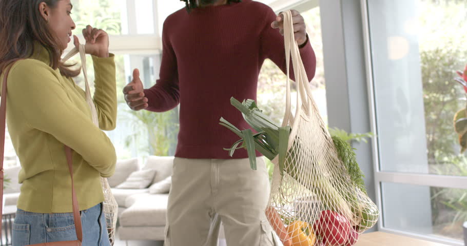 Diverse couple unloading mesh grocery bags onto wooden table near windows, planning dinner prep. Eco-friendly, sustainability, fresh produce, domestic, intimate, modern, natural light