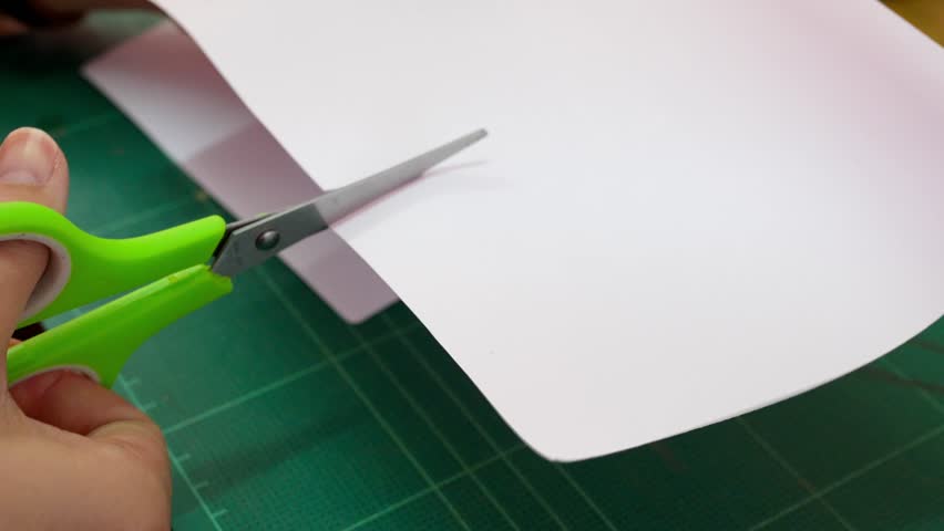 A person is actively using green scissors to cut pink paper on a green cutting mat, highlighting the details of a creative project in a bright indoor environment.