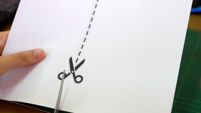 A hand holding scissors is skillfully cutting along a dotted line on a sheet of white paper. The workspace includes a cutting mat and various desk items, emphasizing creativity.