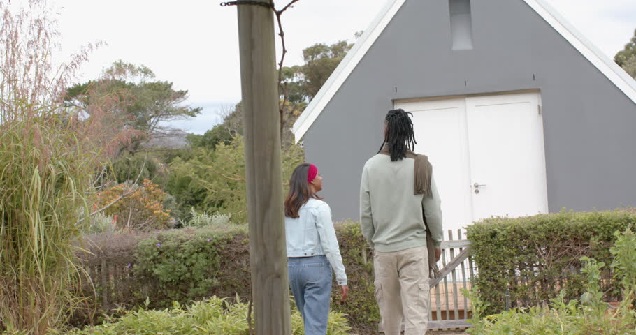 Diverse couple walking along garden path toward cabin spotting white doors, pausing to unlatch gate. Romantic, intimacy, nature, serenity, countryside, leisure, harmony