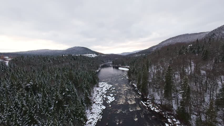 Cinematic dive toward a narrow river running through steep rocky mountains covered in fresh white snow.