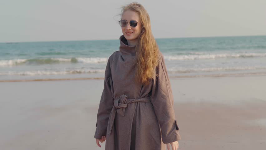 Joyful young woman with long blonde hair wearing sunglasses and coat, spinning around, happily dancing, sandy beach by the sea, concept of expressing happiness, enjoying a beautiful windy day freedom