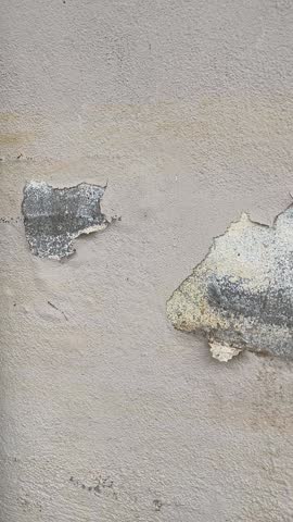 A pan shot from left to right showing a damaged exterior wall with peeling paint and flaking plaster revealing concrete.