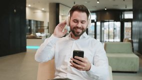 Happy relaxed Caucasian young businessman wearing headphones listening favorite energetic disco music on smartphone. Male entrepreneur in formal relaxing, taking a break sitting in modern office lobby - Powered by Shutterstock - Get 15% off with code: PIKWIZARD15