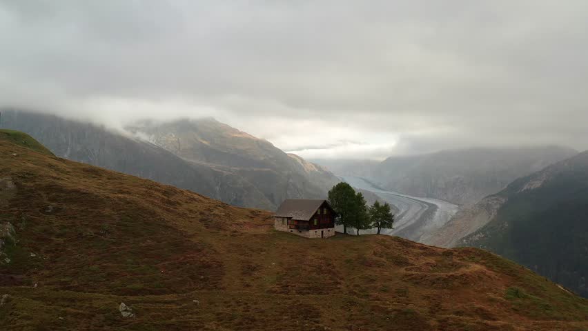 An aerial view of a wooden chalet overlooking the misty Swiss Alps near Belalp, rolling brown hills and the distant Aletsch Glacier beneath clouds