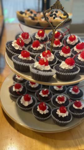 vertical video of mini chocolate cakes with cherries and cream arranged on a tiered stand in a sweet display