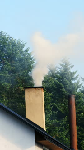 White smoke moves slowly upward from chimney of house. Gentle stream of smoke flows from chimney and fills calm air above house. Vertical shot