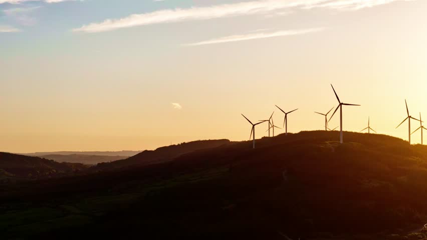 Several wind turbines are spinning atop a grassy hill during the late afternoon. The sun is setting, casting a warm, orange glow over the landscape.