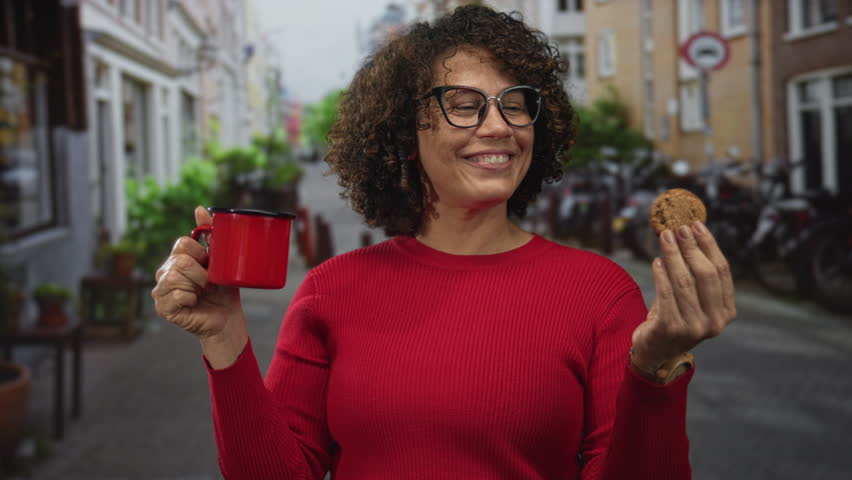 Woman holds red mug and chocolate chip cookie while smiling on cobblestone city street; happiness contentment.