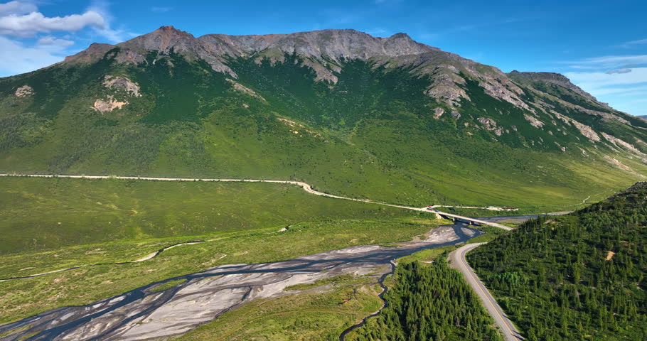 Mountainous landscape crossed by the river and highway with bridge. Drone footage over the natural scenery of Alaska, USA on sunny day.
