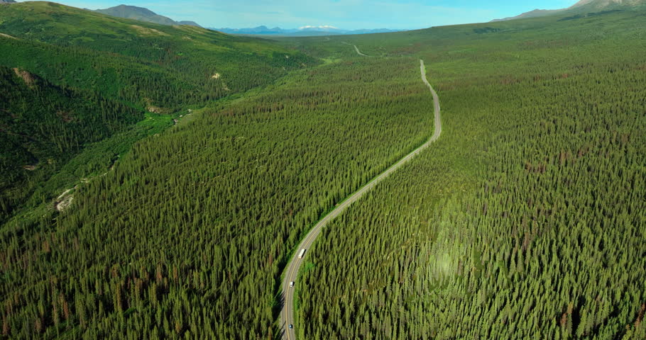 A bus and a car going by the long highway crossing the valley covered with vast evergreen woods. Flight over the wilderness in Alaska, USA on sunny day.