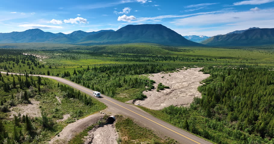 White lonely campervan moving by the highway in the wilderness of Alaska. Aerial perspective on the vast valley with mountain range at backdrop.