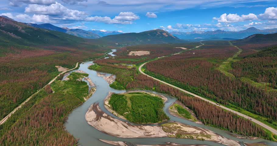 Footage over the branching river flowing in the vast valley in Alaska wilderness. Cars go by the highway crossing the scenery towards the mountains at backdrop.
