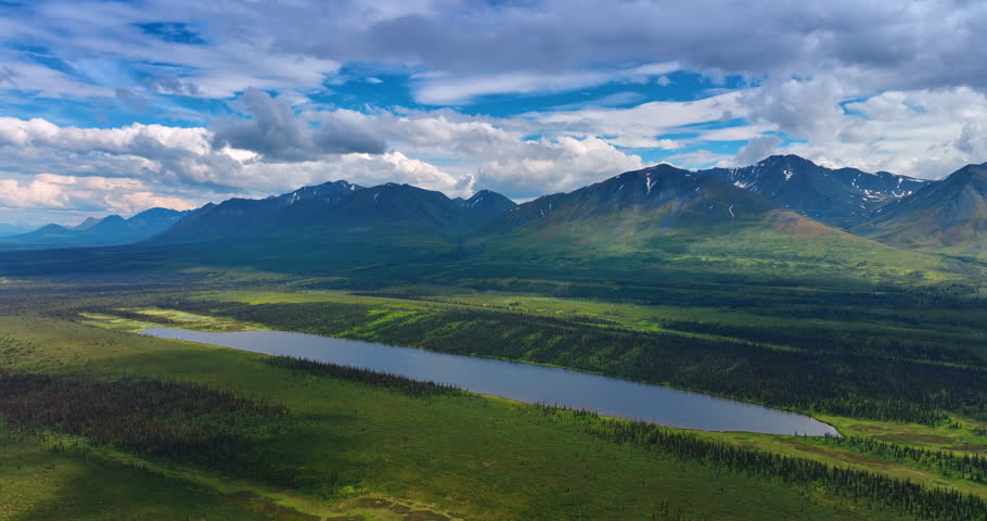 Approaching a long pond among the green meadows. Fluffy white clouds hang over the mountains at backdrop. Alaska aerial.
