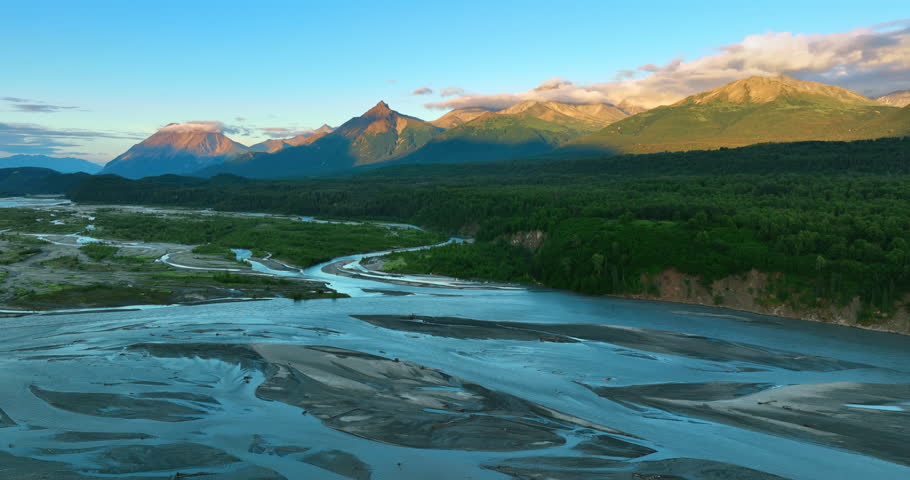 Shallow river flowing the beautiful valley. Setting sun lights the mountain tops partially covered with pink clouds. Alaska wild nature.